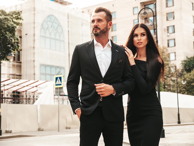 Hermosa mujer de moda y su novio elegante guapo en traje Modelo morena sexy en vestido de noche negro Pareja de moda posando en la calle en Europa Hombre brutal y su mujer al aire libre