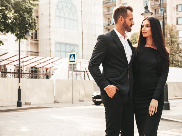 Hermosa mujer de moda y su novio elegante guapo en traje Modelo morena sexy en vestido de noche negro Pareja de moda posando en la calle en Europa Hombre brutal y su mujer al aire libre