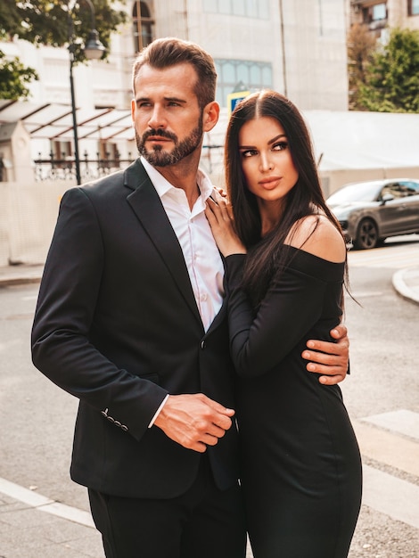 Hermosa mujer de moda y su novio elegante guapo en traje Modelo morena sexy en vestido de noche negro Pareja de moda posando en la calle en Europa Hombre brutal y su mujer al aire libre