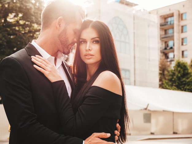 Hermosa mujer de moda y su novio elegante guapo en traje Modelo morena sexy en vestido de noche negro Pareja de moda posando en la calle en Europa Hombre brutal y su mujer al aire libre