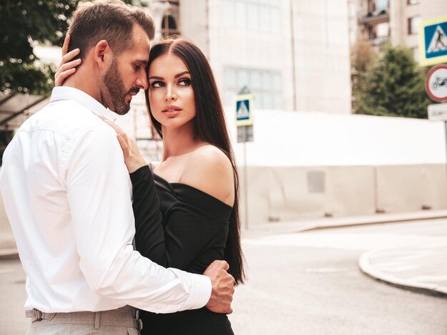 Hermosa mujer de moda y su novio elegante guapo en traje Modelo morena sexy en vestido de noche negro Pareja de moda posando en la calle en Europa Hombre brutal y su mujer al aire libre