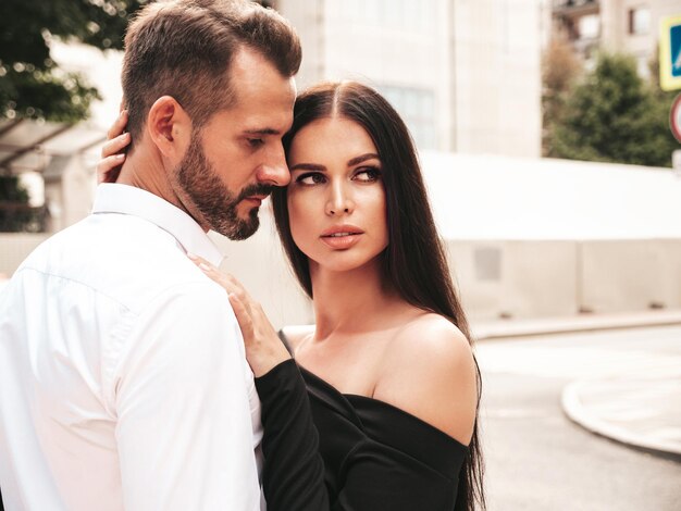 Hermosa mujer de moda y su novio elegante guapo en traje Modelo morena sexy en vestido de noche negro Pareja de moda posando en la calle en Europa Hombre brutal y su mujer al aire libre