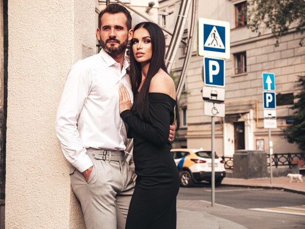 Hermosa mujer de moda y su novio elegante guapo en traje Modelo morena sexy en vestido de noche negro Pareja de moda posando en la calle en Europa Hombre brutal y su mujer al aire libre