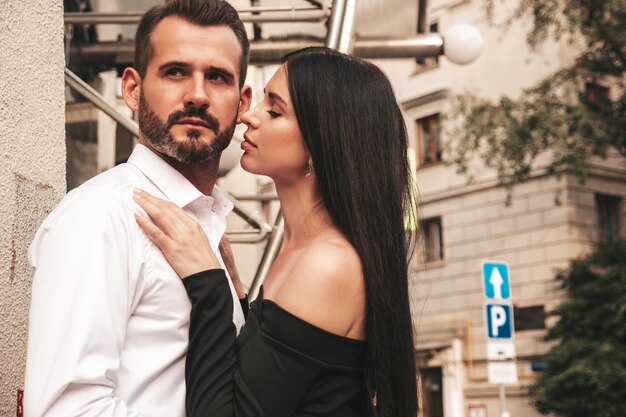 Hermosa mujer de moda y su novio elegante guapo en traje Modelo morena sexy en vestido de noche negro Pareja de moda posando en la calle en Europa Hombre brutal y su mujer al aire libre