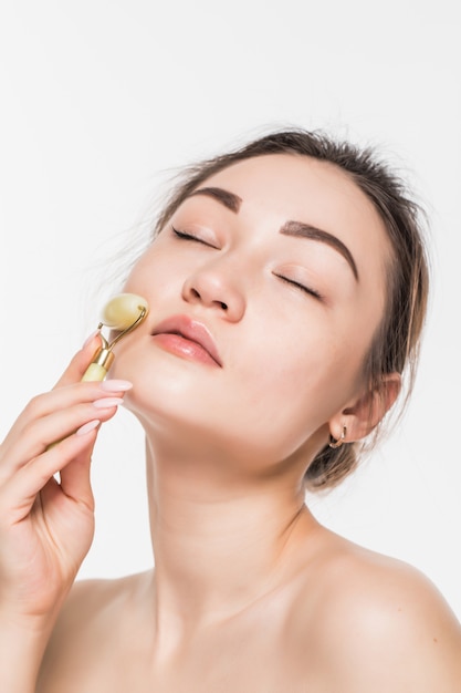 Hermosa mujer modelo con piel limpia fresca y saludable disfrutando de un masaje con un rodillo facial de jade para mejorar la circulación, relajar los músculos y tonificar la piel, aislado en una pared gris con espacio de copia