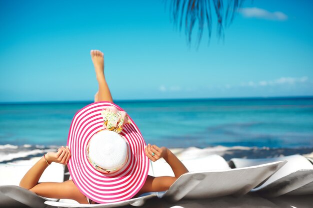 Hermosa mujer modelo tomando el sol en la silla de playa en bikini blanco en colorido sombrero para el sol detrás del océano de agua azul de verano