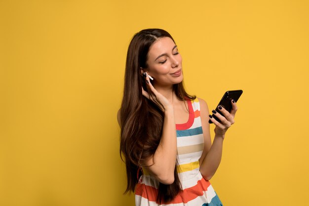 Hermosa mujer moderna con cabello largo oscuro con vestido brillante escuchando música con smartphone sobre pared amarilla.