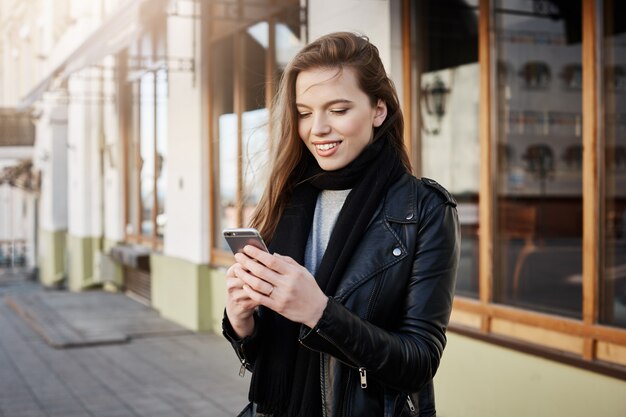 Hermosa mujer moderna en ropa de moda con teléfono inteligente y mirando la pantalla mientras envía mensajes o navega por la red, caminando en la calle