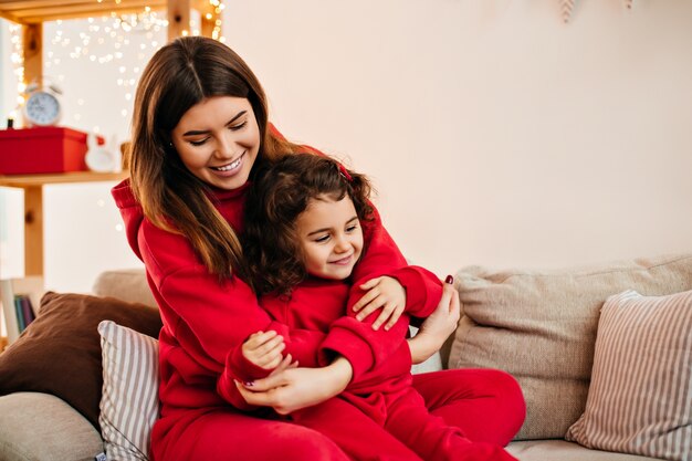 Hermosa mujer morena abrazando al niño con una sonrisa. Mamá joven en traje rojo sentada en el sofá con la pequeña hija.