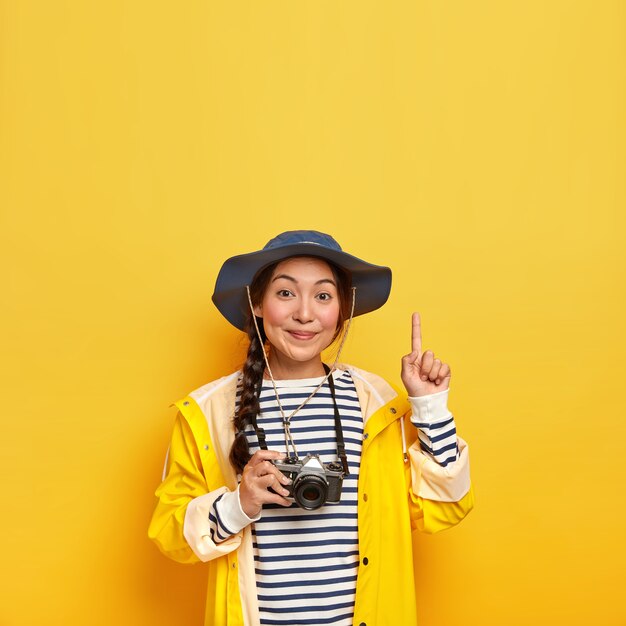 Hermosa mujer morena con apariencia asiática, toma una foto durante el viaje de senderismo con una cámara retro, usa un jersey a rayas, un sombrero y un impermeable, apunta hacia arriba con el dedo índice, aislado sobre una pared amarilla