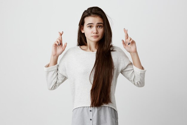 hermosa mujer morena con cabello largo con una mirada excitada, supersticiosa e ingenua, manteniendo los dedos cruzados, esperando buena suerte antes de aprobar el examen. Lenguaje corporal y gestos.