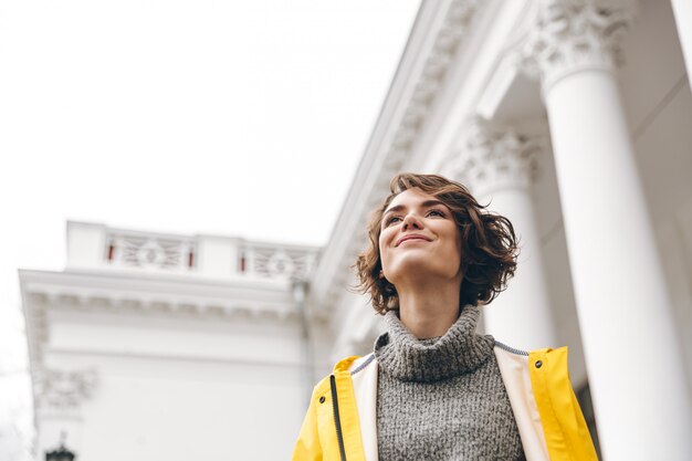 Hermosa mujer morena disfrutando mientras disfruta de puntos de referencia de pie delante del antiguo edificio con amplia sonrisa