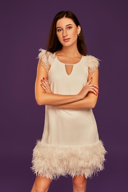 Hermosa mujer morena con un elegante vestido blanco de cóctel posa en el estudio con fondo morado. Foto de estudio de tres cuartos de duración