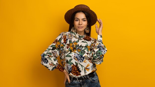 Hermosa mujer morena con estilo atractiva posando aislada sobre fondo de estudio amarillo en ropa de moda de moda blusa impresa y sombrero sonriendo feliz accesorios