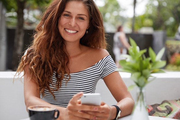 Hermosa mujer morena con expresión alegre y teléfono en la terraza exterior de café