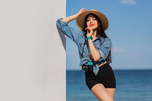 Hermosa mujer morena con figura en pantalones cortos y sombrero de paja posando en la playa.