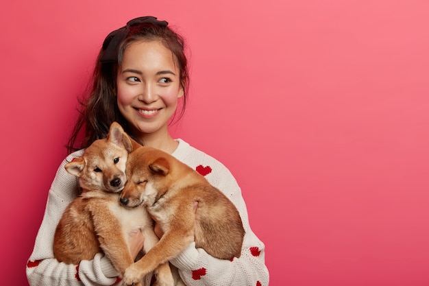 Hermosa mujer morena juega con dos perros shiba inu, mira hacia otro lado, piensa cómo alimentar a las mascotas y enseñar comandos, expresa caricias, aisladas sobre fondo rosa.