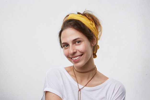 Hermosa mujer morena con pañuelo amarillo en la cabeza, colgante en el cuello y camiseta blanca sonriendo agradablemente con los ojos llenos de felicidad. linda hembra aislada sobre pared blanca en blanco