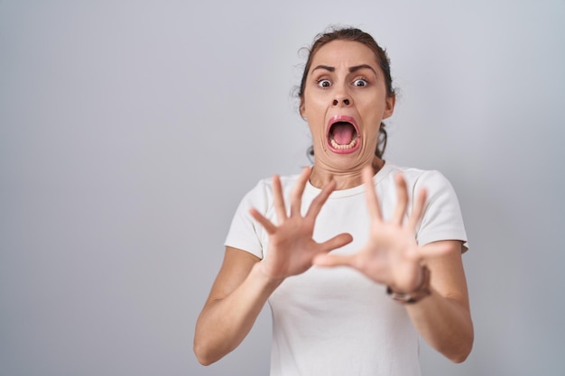 Hermosa mujer morena de pie sobre un fondo aislado asustada y aterrorizada con expresión de miedo detener el gesto con las manos, gritando en estado de shock. concepto de pánico.