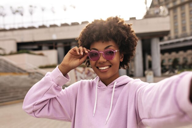 Hermosa mujer morena rizada de piel oscura con elegantes gafas de sol coloridas y una sudadera con capucha rosa sonríe ampliamente y se toma selfie al aire libre