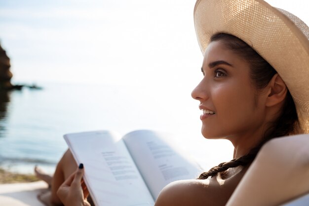 Hermosa mujer morena con sombrero leyendo libro, tumbado en la playa