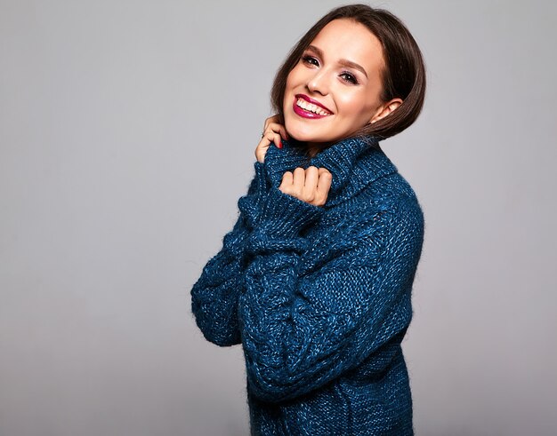 hermosa mujer morena en suéter azul casual con maquillaje brillante y labios rojos sobre gris