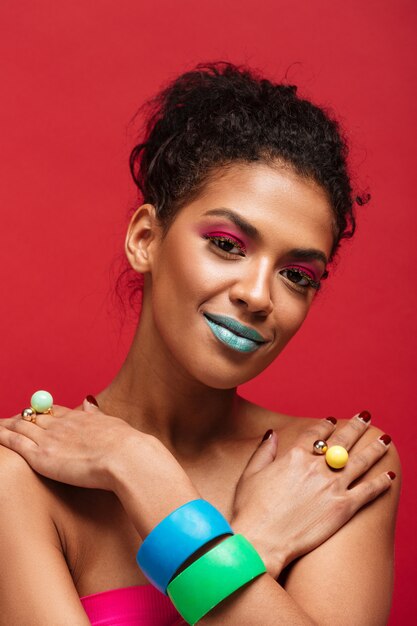 hermosa mujer mulata semidesnuda con maquillaje de moda y accesorios coloridos poniendo las manos cruzadas sobre los hombros, sobre la pared roja