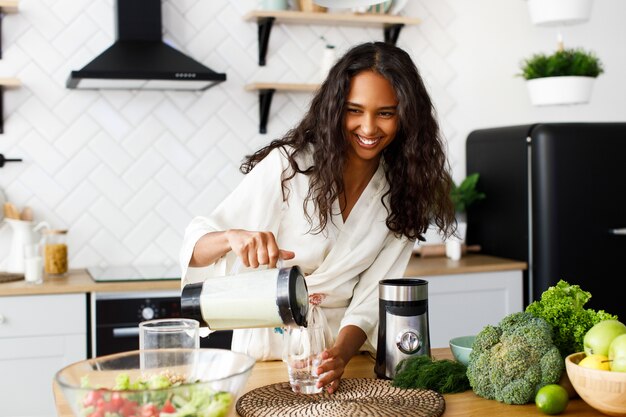 Una hermosa mujer mulata sonrió vertiendo batido verde sobre la cristalería cerca de la mesa con verduras frescas en la cocina moderna blanca vestida con ropa de dormir con el pelo suelto