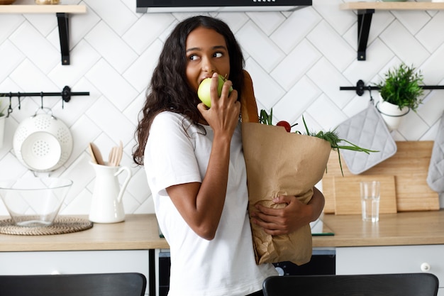 Hermosa mujer mulata sostiene un paquete lleno de comida y come manzana en la moderna cocina blanca