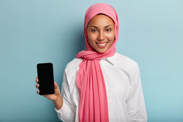 Hermosa mujer musulmana anuncia un dispositivo moderno, sostiene un dispositivo de teléfono inteligente con pantalla en blanco para su anuncio, usa un velo tradicional en la cabeza