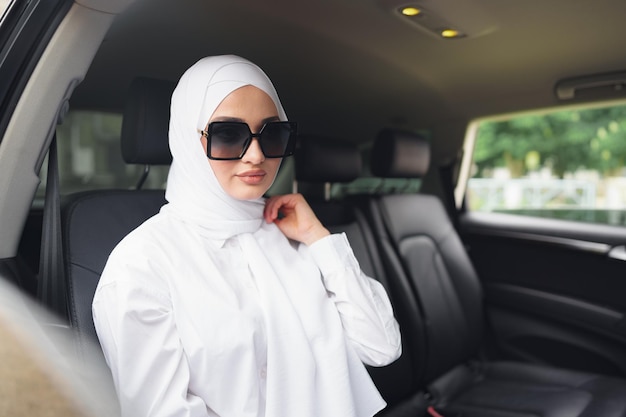 Hermosa mujer musulmana en hijab blanco sentado en el asiento trasero de un coche de lujo