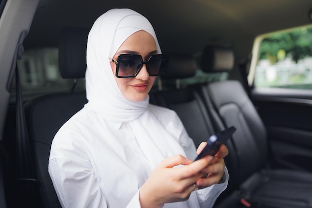 Hermosa mujer musulmana con hiyab blanco sentado en el asiento trasero de un coche y con smartphone