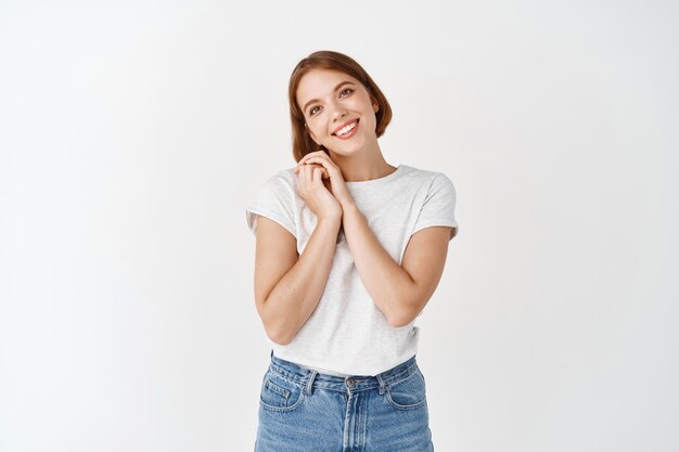 Hermosa mujer natural admira algo, tomados de la mano cerca de la cara y mirando algo lindo, de pie en la pared blanca