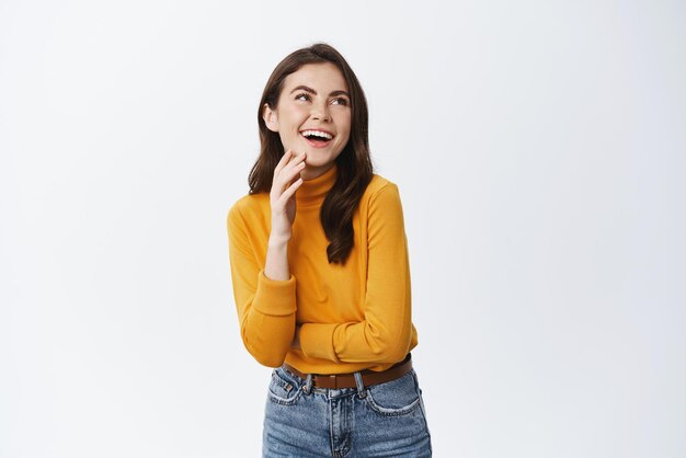 Hermosa mujer natural mirando soñadora y riendo sonriendo feliz de pie en suéter amarillo y jeans contra fondo blanco.