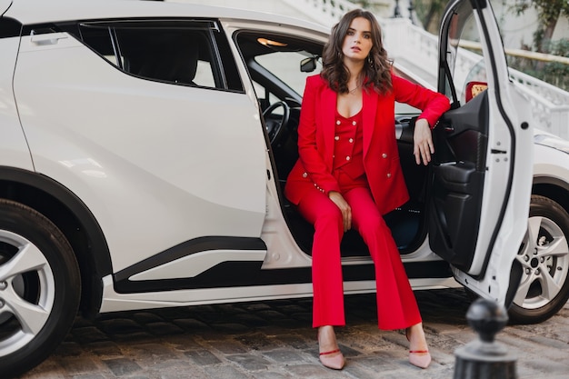 Hermosa mujer de negocios rica sexy en traje rojo posando en coche blanco