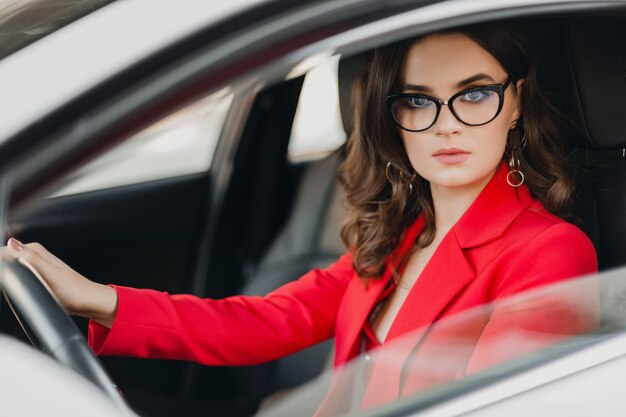 Hermosa mujer de negocios rica sexy en traje rojo sentado en el coche blanco, con gafas