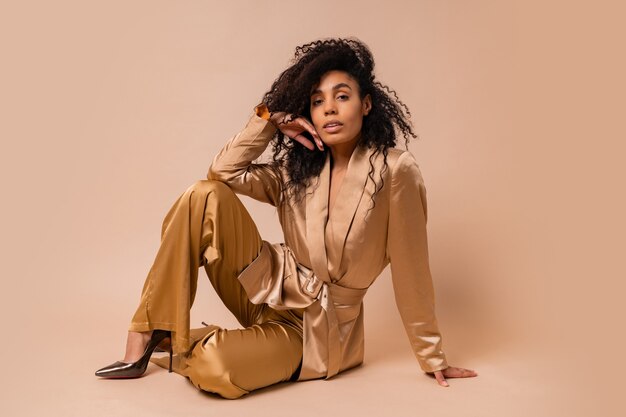 Hermosa mujer negra con hermosos pelos ondulados en un elegante traje de satén dorado posando sobre una pared beige. Look de moda de primavera.