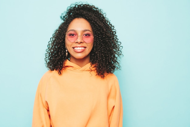 Hermosa mujer negra con peinado afro rizos. Modelo sonriente en sudadera con capucha naranja y ropa de jeans de moda