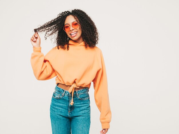 Hermosa mujer negra con peinado afro rizos. Modelo sonriente en sudadera con capucha naranja y ropa de jeans de moda