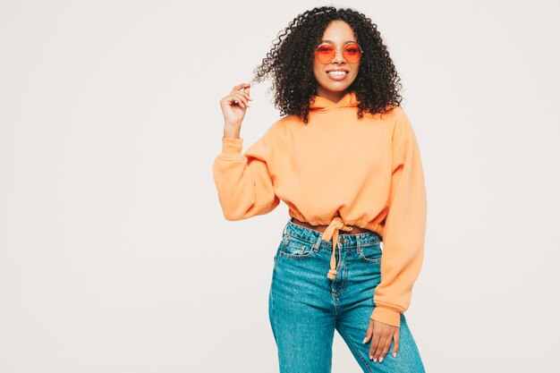 Hermosa mujer negra con peinado afro rizos. Modelo sonriente en sudadera con capucha naranja y ropa de jeans de moda