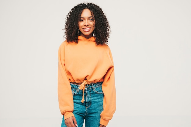 Hermosa mujer negra con peinado afro rizos. Modelo sonriente en sudadera con capucha naranja y ropa de jeans de moda
