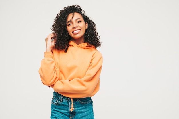 Hermosa mujer negra con peinado afro rizos. Modelo sonriente en sudadera con capucha naranja y ropa de jeans de moda