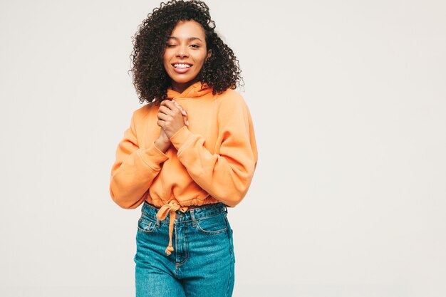 Hermosa mujer negra con peinado afro rizos. Modelo sonriente en sudadera con capucha naranja y ropa de jeans de moda