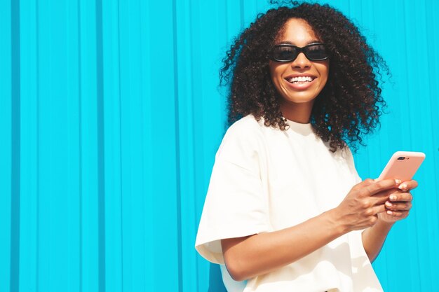 Hermosa mujer negra con peinado de rizos afro Modelo hipster sonriente en camiseta blanca Sexy mujer despreocupada posando en la calle cerca de la pared azul con gafas de sol Mirando la pantalla del teléfono inteligente usando aplicaciones