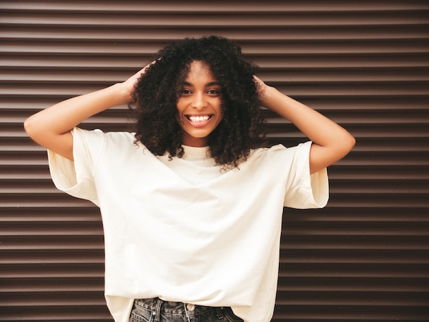 Hermosa mujer negra con peinado de rizos afro Modelo hipster sonriente en camiseta blanca Sexy mujer despreocupada posando en la calle cerca de la pared marrón Alegre y feliz en gafas de sol