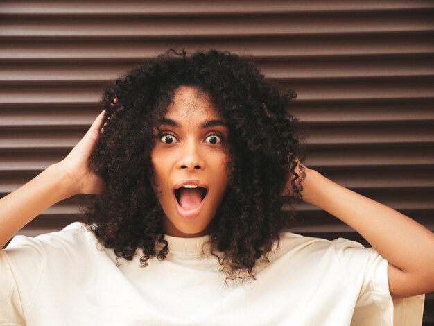 Hermosa mujer negra con peinado de rizos afro Modelo hipster sonriente en camiseta blanca Sexy mujer despreocupada posando en la calle cerca de la pared marrón Alegre y feliz Sorprendida y sorprendida