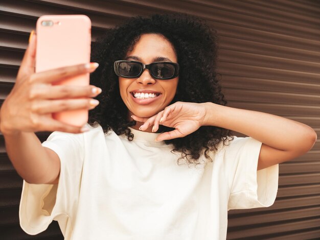 Hermosa mujer negra con peinado de rizos afro Modelo hipster sonriente en camiseta blanca Sexy mujer despreocupada posando en la calle cerca de la pared marrón con gafas de sol Alegre y feliz Tomando selfie