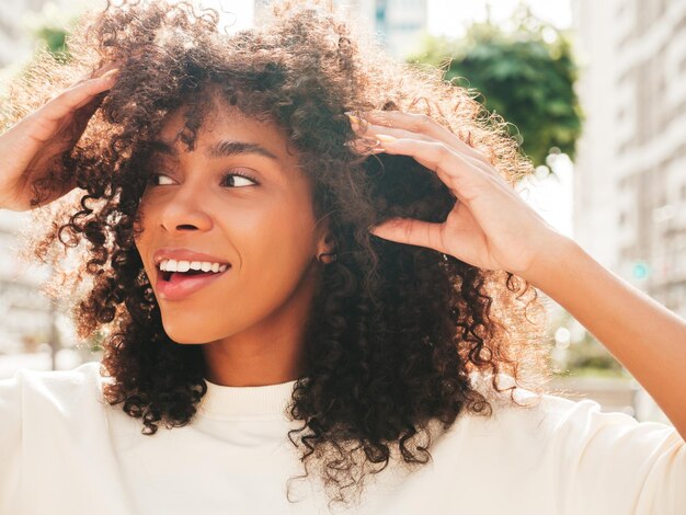 Hermosa mujer negra con peinado de rizos afro Modelo hipster sonriente en camiseta blanca Sexy mujer despreocupada posando en el fondo de la calle Alegre y feliz