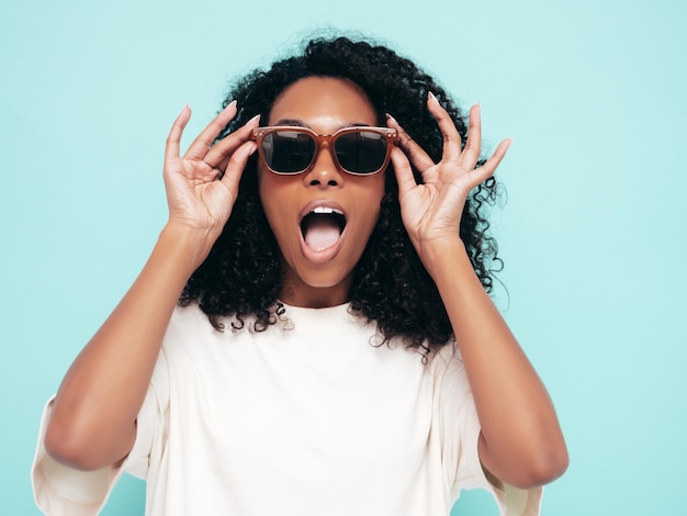 Hermosa mujer negra con peinado de rizos afro Modelo sonriente en ropa de camiseta larga Mujer sexy despreocupada posando junto a la pared azul en el estudio Bronceada y alegre con gafas de sol Sorprendida y sorprendida