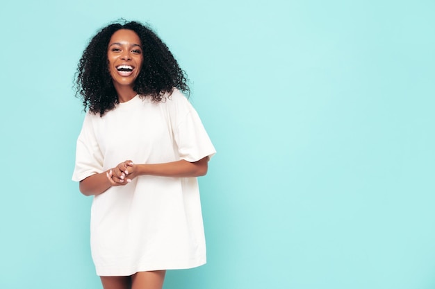 Hermosa mujer negra con peinado de rizos afro Modelo sonriente en ropa de camiseta larga Mujer sexy despreocupada posando junto a la pared azul en el estudio Bronceada y alegre